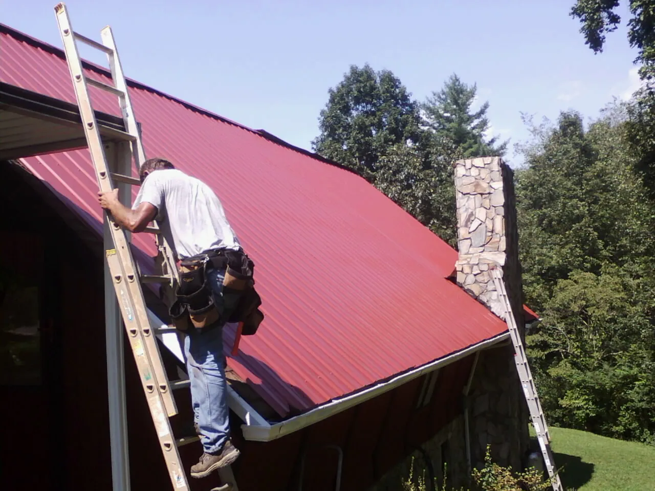 Roofer on a ladder beside a red metal roof and stone chimney — Soffit & Fascia Repair by Roofing Brothers in Westlake Village